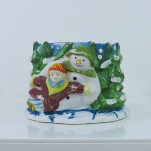 Könnte beinhalten: Ein keramischer Weihnachtskerzenhalter mit einem Schneemann und einem Kind. Der Schneemann trägt einen grünen Hut und Schal, das Kind einen roten Mantel. Der Hintergrund zeigt grüne Bäume und blauen Himmel mit einer schneebedeckten Basis. Der Kerzenhalter ist etwa 10 cm hoch.