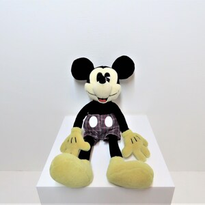 May include: A black and yellow plush Mickey Mouse toy wearing red and black plaid shorts with white polka dots. The toy is sitting on a white surface.