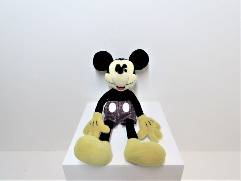 May include: A black and yellow plush Mickey Mouse toy wearing red and black plaid shorts with white polka dots. The toy is sitting on a white surface.