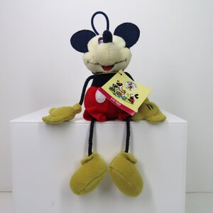 May include: A plush Mickey Mouse toy with long legs and a red and white outfit. The toy is sitting on a white surface and has a tag that says "GUND" on it.