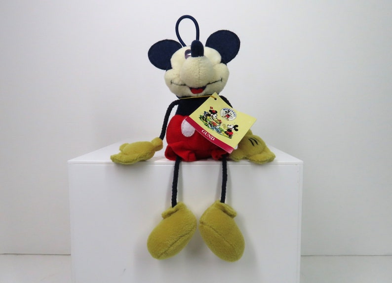 May include: A plush Mickey Mouse toy with long legs and a red and white outfit. The toy is sitting on a white surface and has a tag that says "GUND" on it.
