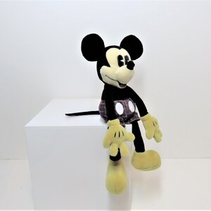 May include: A black and yellow plush Mickey Mouse toy wearing red and black plaid shorts. The toy is sitting on a white surface.