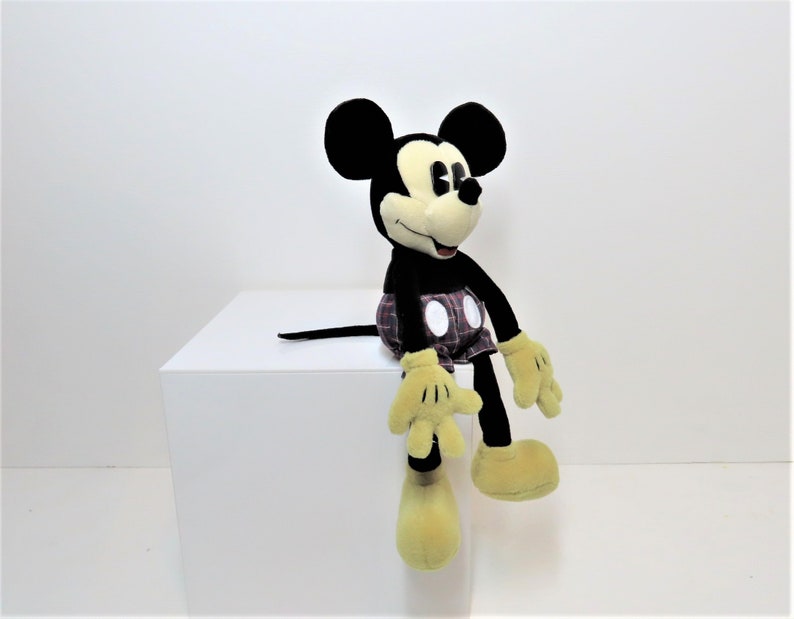 May include: A black and yellow plush Mickey Mouse toy wearing red and black plaid shorts. The toy is sitting on a white surface.