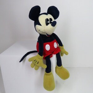 May include: A plush Mickey Mouse toy sitting on a white surface. The toy is wearing red shorts with white polka dots and has black ears and a black nose. The toy has yellow hands and feet.
