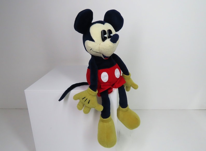 May include: A plush Mickey Mouse toy sitting on a white surface. The toy is wearing red shorts with white polka dots and has black ears and a black nose. The toy has yellow hands and feet.