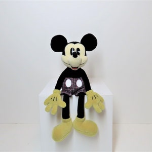 May include: A black and yellow plush Mickey Mouse toy wearing red and black plaid shorts with white polka dots. The toy is sitting on a white surface.