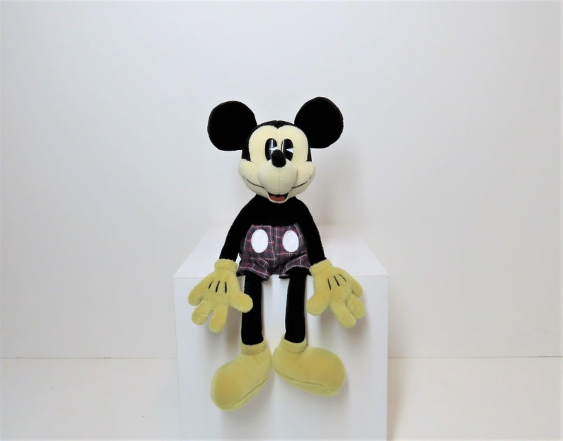 May include: A black and yellow plush Mickey Mouse toy wearing red and black plaid shorts with white polka dots. The toy is sitting on a white surface.