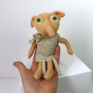 May include: A knitted doll of a fictional character with large green eyes, a long nose, and large ears. The doll is wearing a tan-colored outfit with a matching belt. The doll is a handmade item.