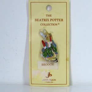 Może przedstawiać: Broszka z kolekcji Beatrix Potter przedstawiająca Królika Piotrusia w niebieskiej kurtce trzymającego marchewkę. Broszka znajduje się na żółtej kartce z tekstem "The Beatrix Potter Collection" i "Brooch".