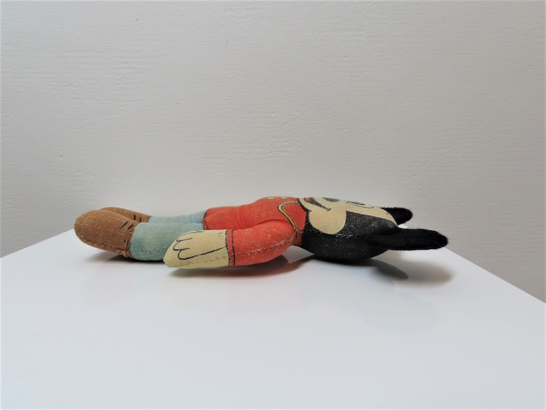 K&ouml;nnte beinhalten: Ein Vintage-Pl&uuml;schtier von Mickey Mouse, das auf der Seite liegt. Das Spielzeug ist aus Stoff und hat ein rotes Hemd, eine blaue Hose und schwarze Ohren. Das Spielzeug ist in gutem Zustand, weist aber Gebrauchsspuren auf.
