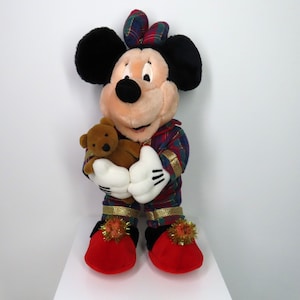 May include: A plush Mickey Mouse toy wearing red and green plaid pajamas and holding a brown teddy bear. The toy has a red bow on its head and is wearing red slippers with gold trim and pom-poms.