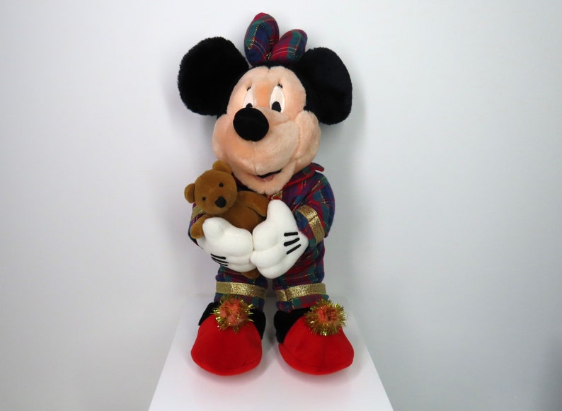 May include: A plush Mickey Mouse toy wearing red and green plaid pajamas and holding a brown teddy bear. The toy has a red bow on its head and is wearing red slippers with gold trim and pom-poms.