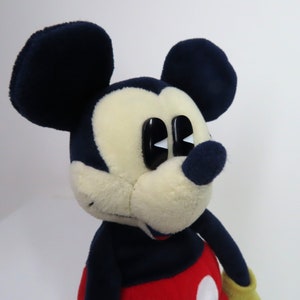 May include: A plush Mickey Mouse toy with black ears, a black nose, and large white eyes. The toy is wearing a red and yellow outfit.
