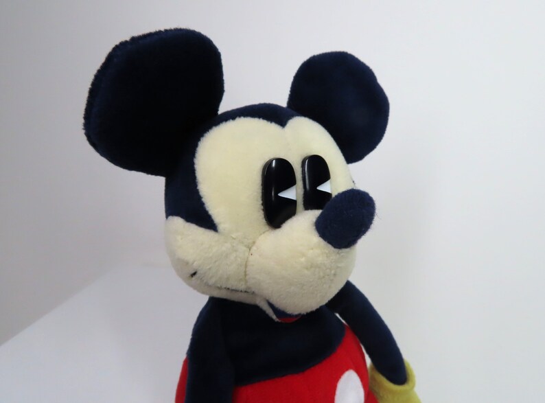 May include: A plush Mickey Mouse toy with black ears, a black nose, and large white eyes. The toy is wearing a red and yellow outfit.