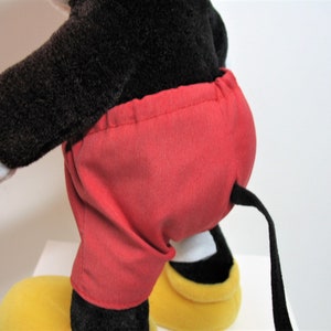 May include: A plush Mickey Mouse toy with black fur, red shorts, and a black tail. The toy is standing on yellow shoes.