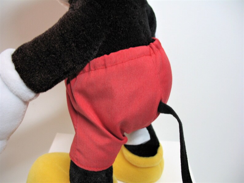 May include: A plush Mickey Mouse toy with black fur, red shorts, and a black tail. The toy is standing on yellow shoes.
