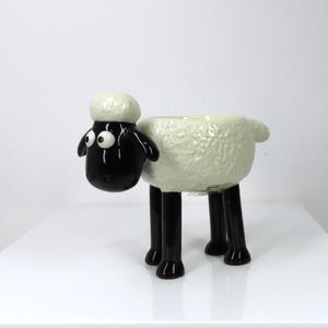 Puede incluir: Una maceta de cerámica con forma de oveja. La oveja tiene cara, orejas y patas negras, y cuerpo y cabeza blancos. La maceta tiene un cuerpo texturizado y está diseñada para contener pequeñas plantas o flores.