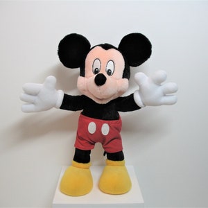 May include: A plush Mickey Mouse toy with black ears, a black body, red shorts with white polka dots, and yellow shoes. The toy is standing with its arms outstretched.