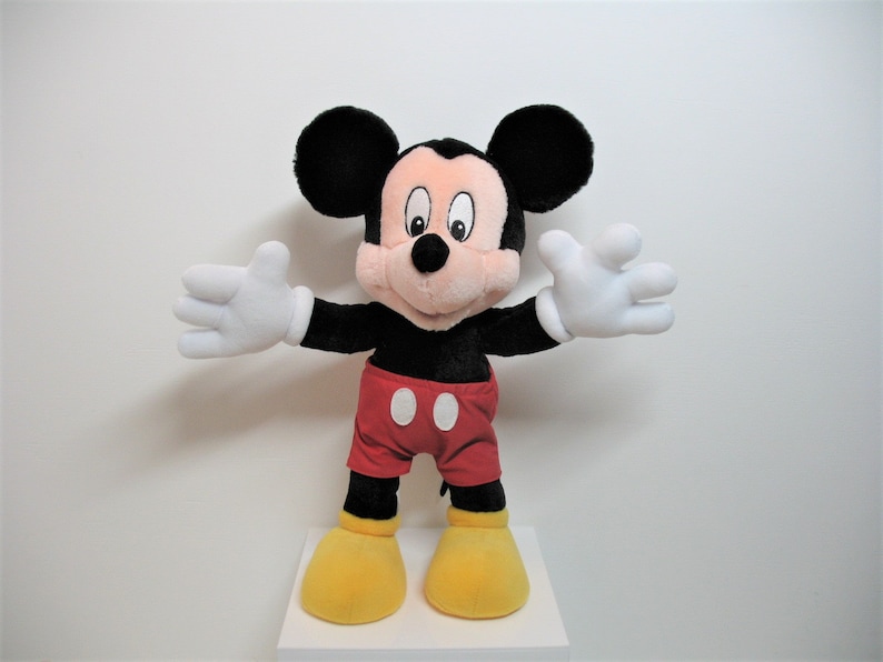 May include: A plush Mickey Mouse toy with black ears, a black body, red shorts with white polka dots, and yellow shoes. The toy is standing with its arms outstretched.