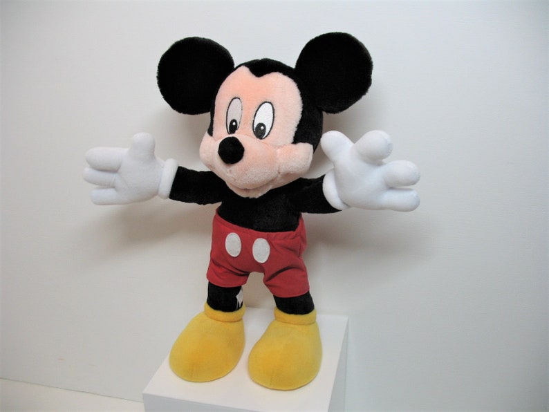 May include: A plush Mickey Mouse toy with black ears, a red and white polka dot shirt, and yellow shoes. The toy is standing on a white surface.