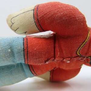 K&ouml;nnte beinhalten: Eine Vintage-Stoffpuppe aus rotem, blauem und schwarzem Stoff. Die Puppe tr&auml;gt ein rot-gelb gestreiftes Hemd und eine blaue Hose. Das Gesicht der Puppe ist wei&szlig; mit schwarzen Augen und einem schwarzen Mund.