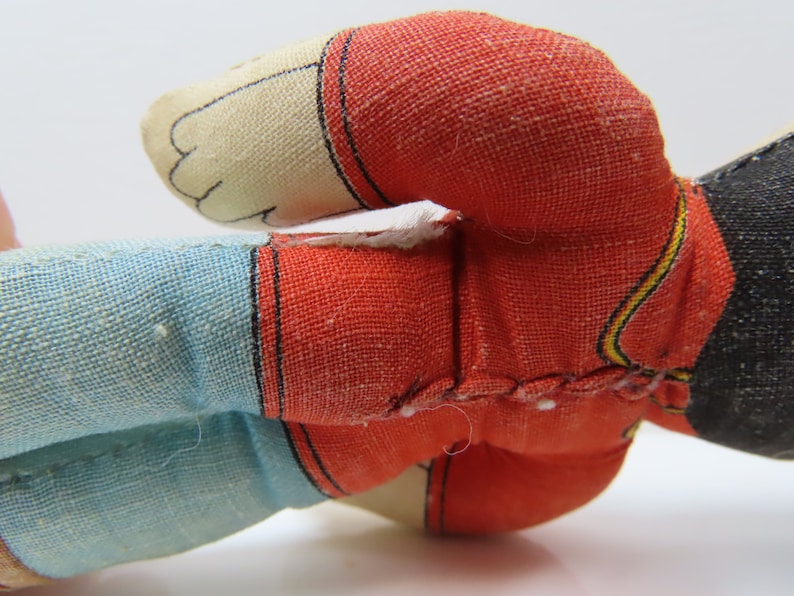 K&ouml;nnte beinhalten: Eine Vintage-Stoffpuppe aus rotem, blauem und schwarzem Stoff. Die Puppe tr&auml;gt ein rot-gelb gestreiftes Hemd und eine blaue Hose. Das Gesicht der Puppe ist wei&szlig; mit schwarzen Augen und einem schwarzen Mund.
