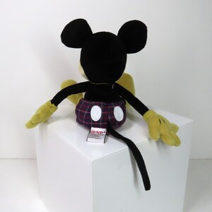 Op de afbeelding: Een zwart en geel pluche Mickey Mouse speelgoed dat op een wit oppervlak zit. Mickey draagt een rood en blauw geruit broekje met witte stippen. Het speelgoed heeft een zwarte staart en een "GUND"-label dat aan de achterkant is bevestigd.
