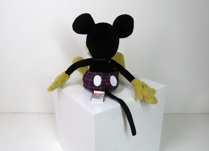 Op de afbeelding: Een zwart en geel pluche Mickey Mouse speelgoed dat op een wit oppervlak zit. Mickey draagt een rood en blauw geruit broekje met witte stippen. Het speelgoed heeft een zwarte staart en een "GUND"-label dat aan de achterkant is bevestigd.