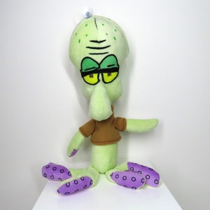 May include: Green plush toy of Squidward Tentacles from Spongebob Squarepants. He is wearing a brown shirt and has purple spots on his tentacles.