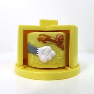 May include: A yellow plastic toy with a spinning cylinder featuring Tom and Jerry characters. Tom is shown with his paw outstretched and Jerry is running away from him.