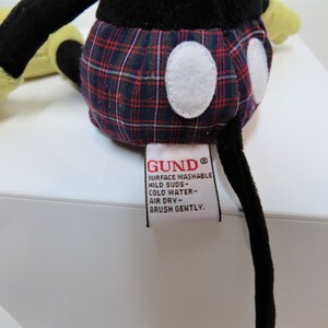 May include: A black and yellow plush Mickey Mouse toy wearing a red, blue, and white plaid shirt. The toy has a white label on its back that reads "GUND&reg; SURFACE WASHABLE MILD SUDS - COLD WATER - AIR DRY - BRUSH GENTLY."