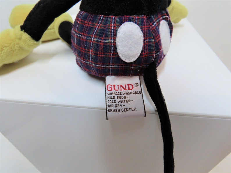 May include: A black and yellow plush Mickey Mouse toy wearing a red, blue, and white plaid shirt. The toy has a white label on its back that reads "GUND&reg; SURFACE WASHABLE MILD SUDS - COLD WATER - AIR DRY - BRUSH GENTLY."