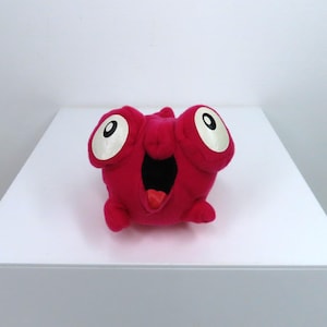 Puede incluir: Un peluche con forma de criatura hecho de tela magenta vibrante, con dos grandes ojos blancos con pupilas negras y una boca muy abierta que revela un interior oscuro y una pequeña lengua roja. El personaje de peluche tiene una forma redondeada.
