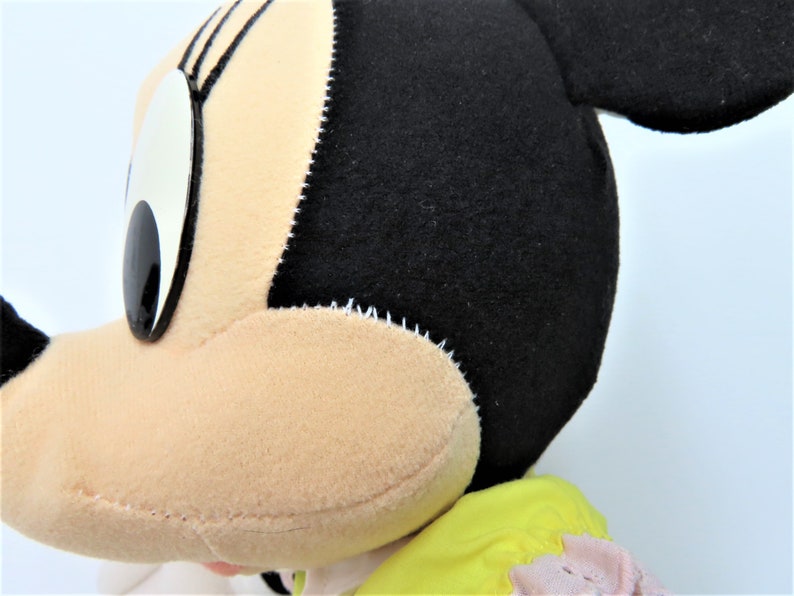 Puede incluir: Primer plano de un rat&oacute;n de peluche con orejas negras, ojos grandes y cara rosa. El rat&oacute;n lleva un vestido amarillo y rosa.