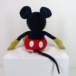 May include: A plush Mickey Mouse toy sitting with its tail behind it. The toy is red, black, yellow, and white. The toy has a red and white polka dot shorts and a black tail.
