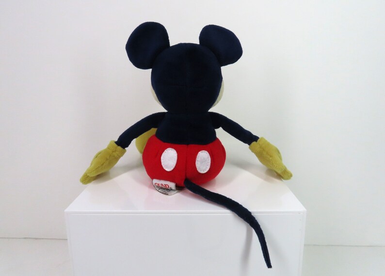 May include: A plush Mickey Mouse toy sitting with its tail behind it. The toy is red, black, yellow, and white. The toy has a red and white polka dot shorts and a black tail.