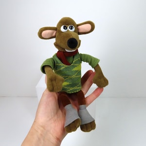 Puede incluir: Un ratón de peluche marrón que lleva un suéter verde con una bufanda roja. El ratón tiene ojos marrones grandes y una amplia sonrisa.