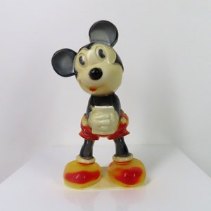 May include: A vintage plastic Mickey Mouse figurine with black ears, a black nose, and red shorts. The figurine is standing on a white surface.