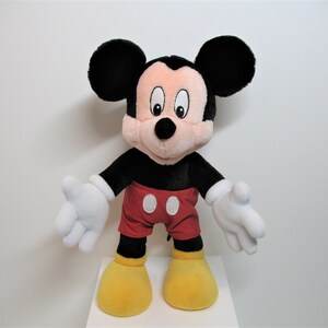 May include: A plush Mickey Mouse toy with black ears, a black body, red shorts with a white circle, and yellow shoes. The toy is standing on a white surface.