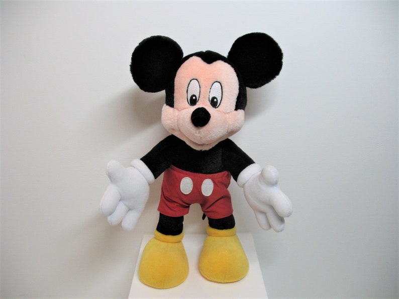 May include: A plush Mickey Mouse toy with black ears, a black body, red shorts with a white circle, and yellow shoes. The toy is standing on a white surface.