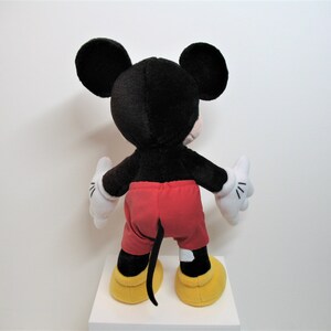 May include: A black and red plush Mickey Mouse toy with yellow shoes. The toy is standing on a white surface.