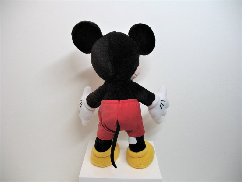 May include: A black and red plush Mickey Mouse toy with yellow shoes. The toy is standing on a white surface.