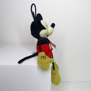 May include: A plush Mickey Mouse toy sitting on a white surface. Mickey is wearing his signature red shorts with a yellow button and black shoes. He has a black nose and large, round ears.