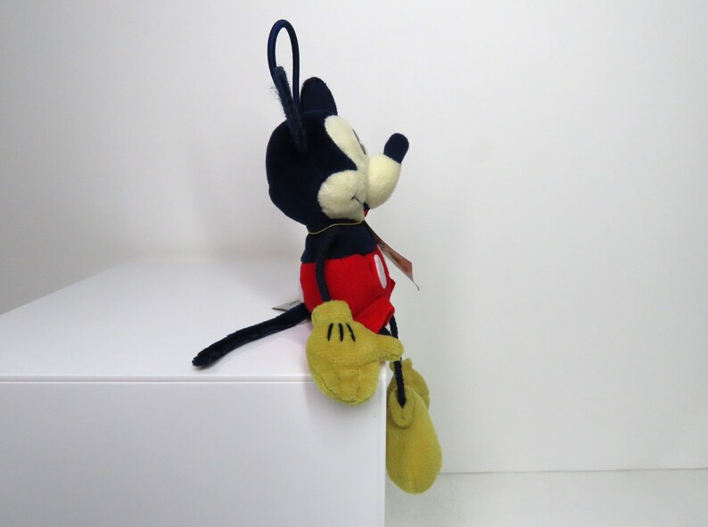 May include: A plush Mickey Mouse toy sitting on a white surface. Mickey is wearing his signature red shorts with a yellow button and black shoes. He has a black nose and large, round ears.