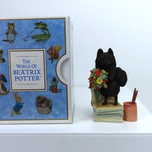 May include: A collectible figurine of a black cat holding a bouquet of flowers, standing on a stack of books, next to a small orange pot with red sticks. A box with illustrations of Beatrix Potter characters is on the left.