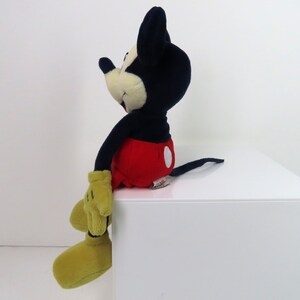 May include: A plush Mickey Mouse toy sitting on a white surface. Mickey is wearing his signature red shorts with a white circle, yellow shoes, and black and white gloves. He has a black nose and a big smile.