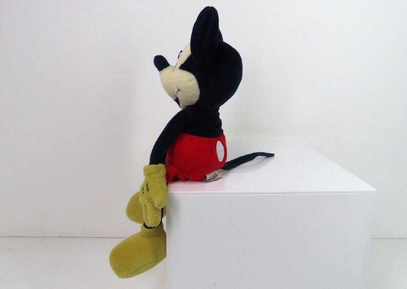 May include: A plush Mickey Mouse toy sitting on a white surface. Mickey is wearing his signature red shorts with a white circle, yellow shoes, and black and white gloves. He has a black nose and a big smile.