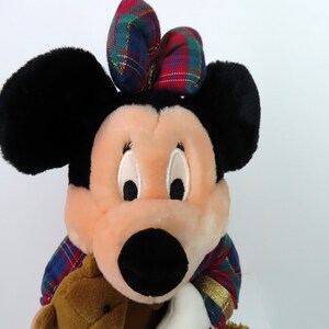 May include: A plush Minnie Mouse toy wearing a red, green, and blue plaid bow and a matching plaid scarf. She is holding a brown teddy bear.