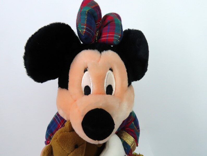 May include: A plush Minnie Mouse toy wearing a red, green, and blue plaid bow and a matching plaid scarf. She is holding a brown teddy bear.