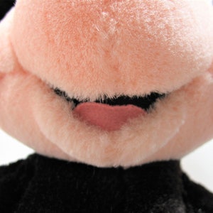 May include: Close-up of a plush toy's face. The toy has a pink nose and mouth, with a black mouth and a pink tongue.